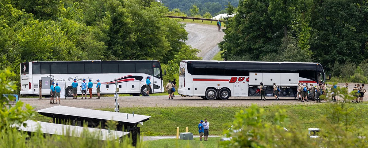 Two white charter buses parked along a gravel road in a lush, wooded area, with Scouts in uniform gathering and unloading luggage from open cargo bays.