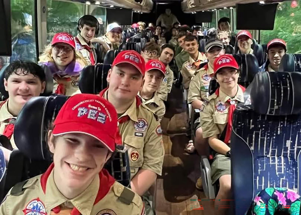 A group of Scouts wearing tan uniforms, red neckerchiefs, and matching red baseball caps smiling while seated inside a charter bus.