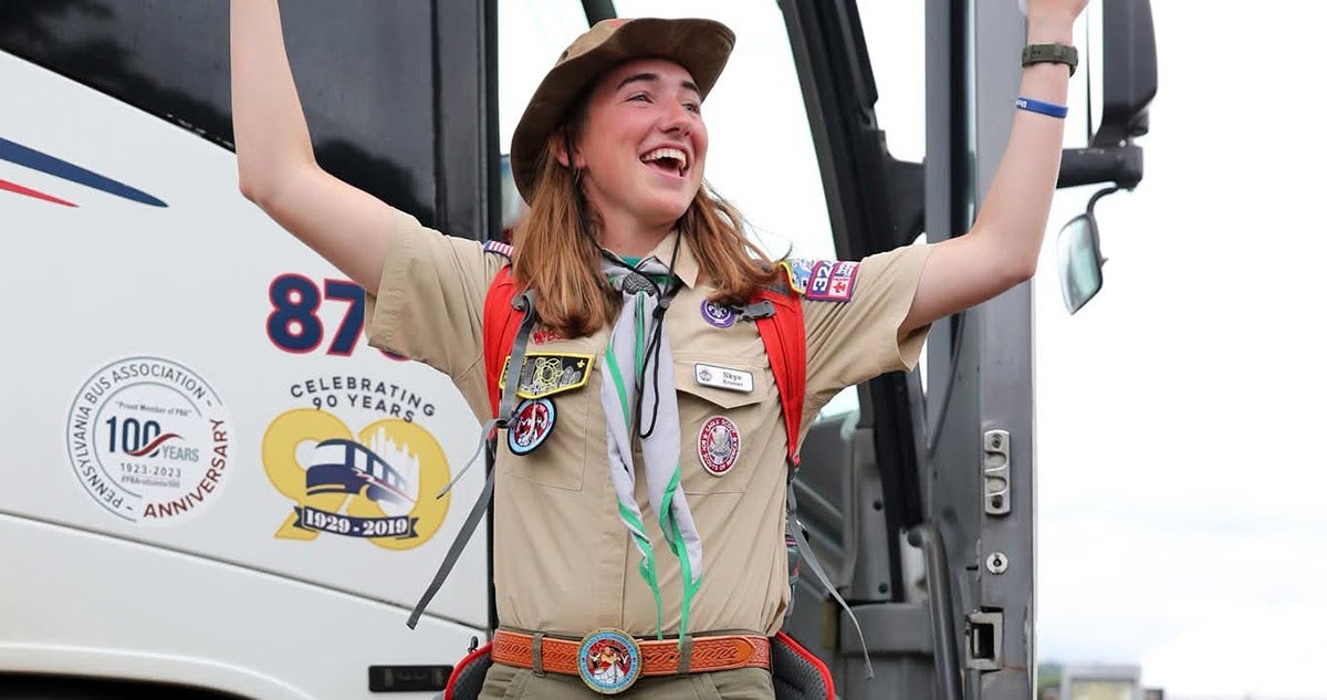 A joyful Scout wearing a full tan uniform and hat raises her arms in celebration while standing outside the open door of a charter bus.