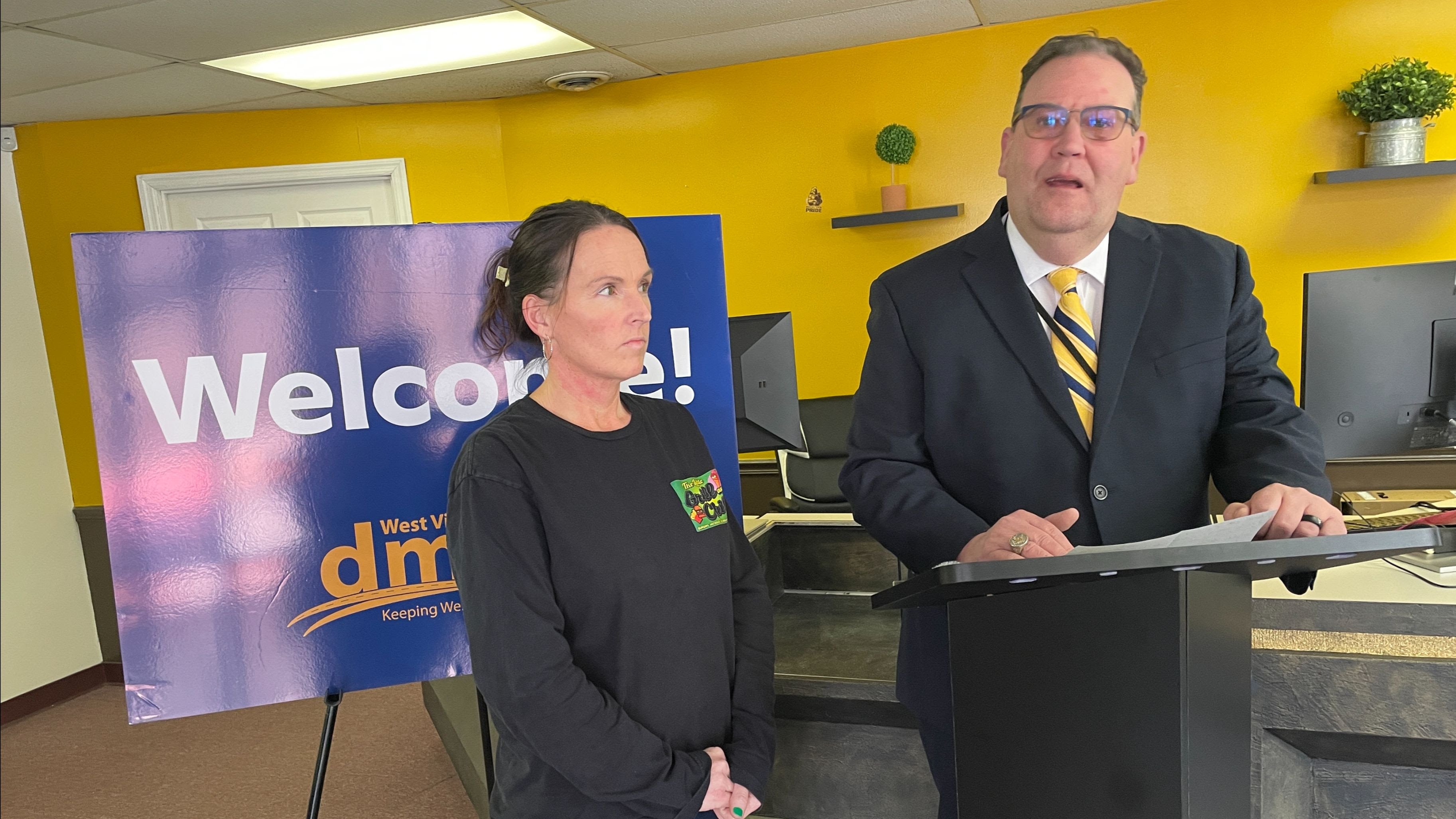 A West Virginia DMV official speaks at a podium next to a woman and a blue DMV welcome sign.