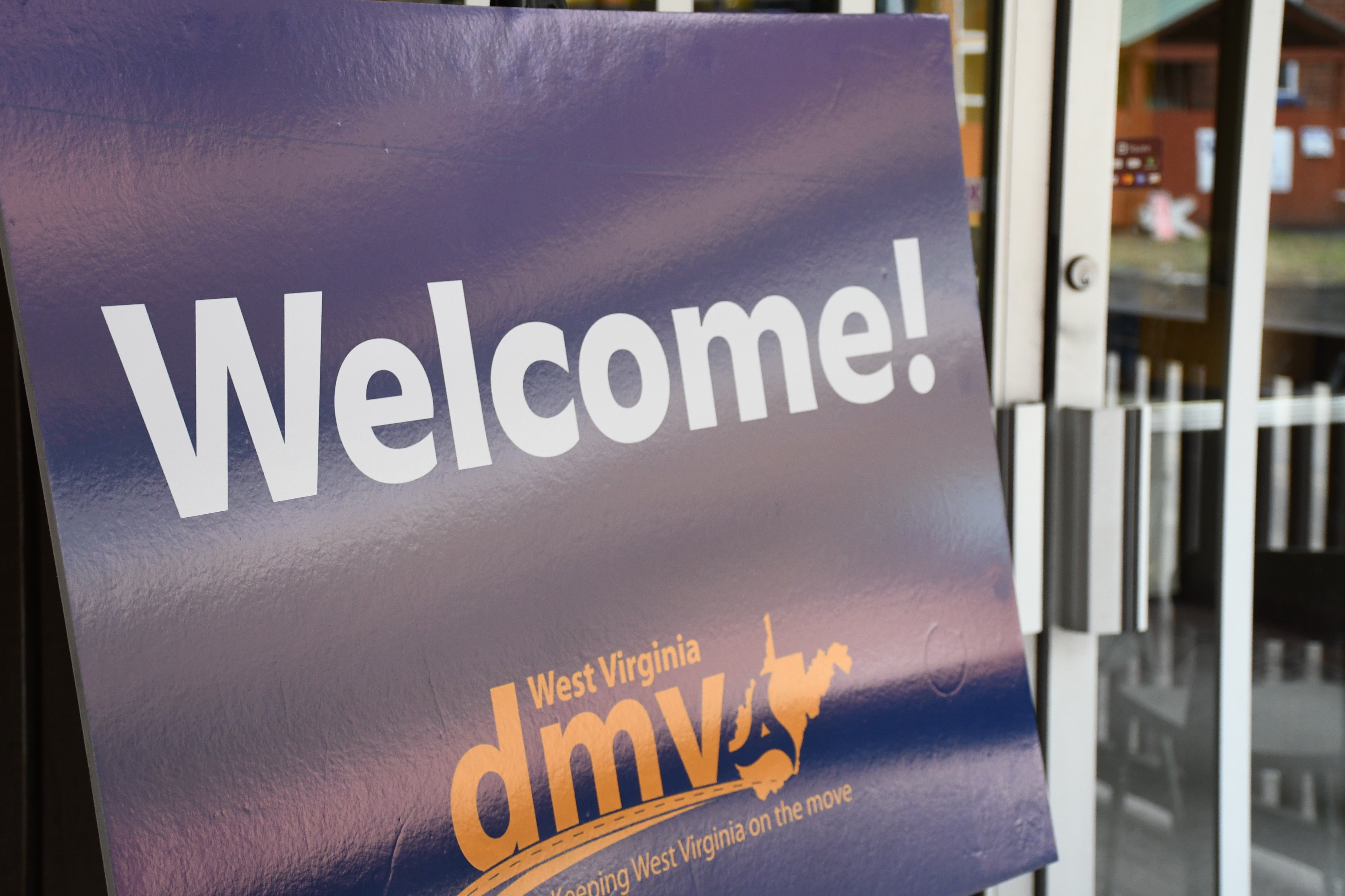 A close-up of a blue West Virginia DMV sign that reads 'Welcome!' positioned next to a glass entrance door.