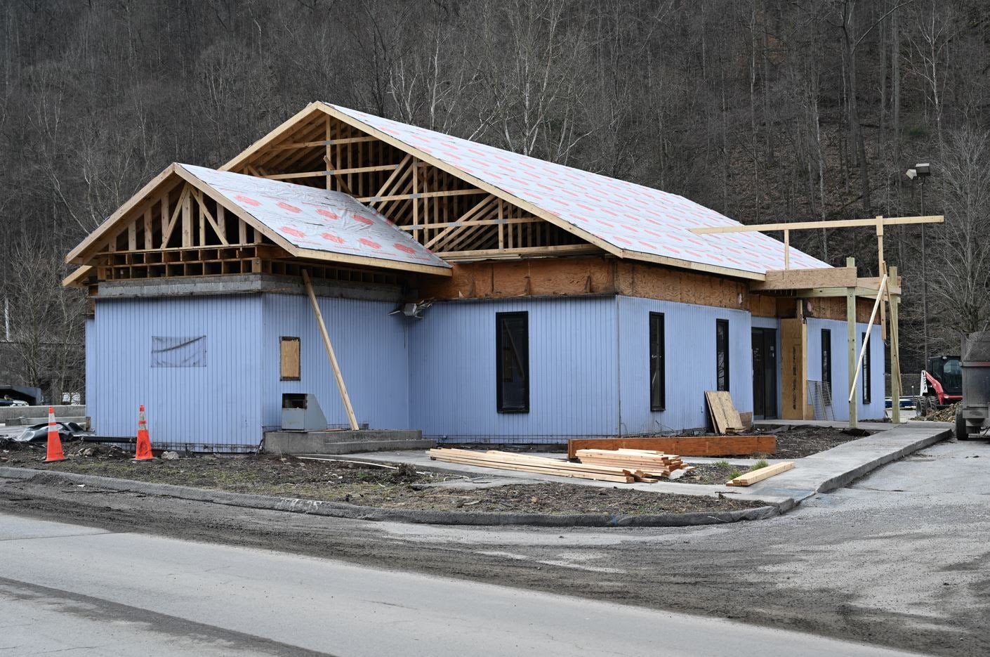 A single-story light blue building with a new wooden roof truss structure under construction, with building materials on the ground and a wooded hillside background.