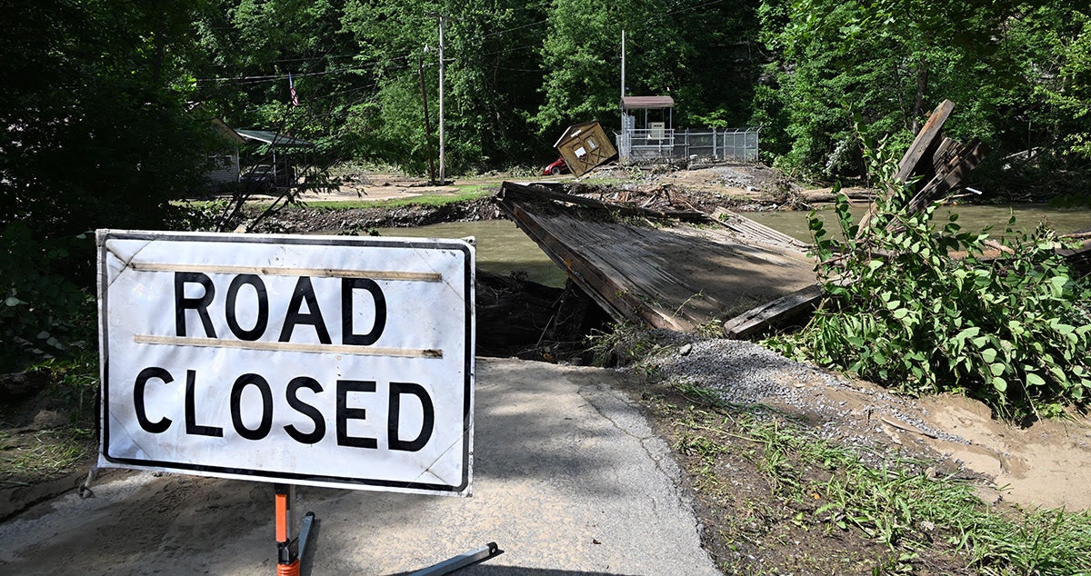 A large 'ROAD CLOSED' sign stands in the foreground, blocking a road where a wooden bridge has collapsed into a river due to severe flood damage