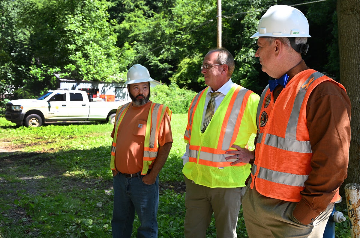 Three men wearing hard hats and high-visibility safety vests stand in a grassy area discussing flood recovery efforts, with a white WVDOH truck in the background