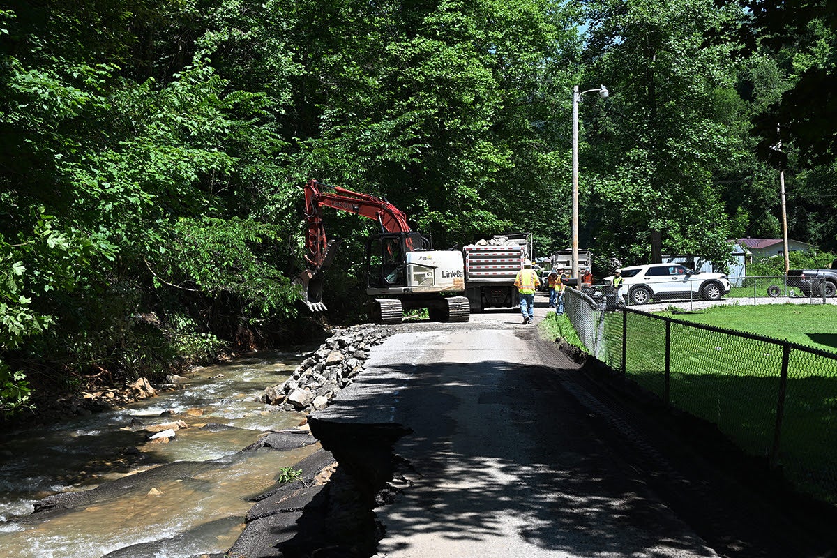 An excavator and work crew reinforce a heavily eroded, washed-out asphalt road with stone riprap next to a flowing creek