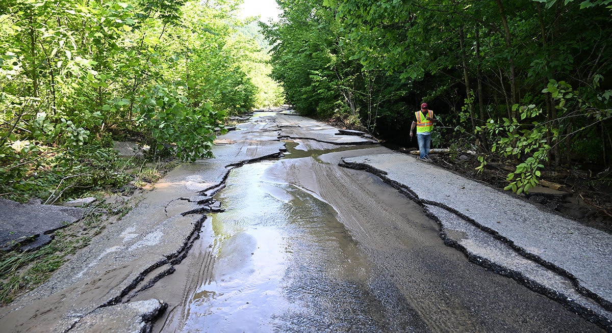 A severely fractured and buckled asphalt road covered in mud and standing water from flood damage, with a highway worker walking along the edge to survey the destruction