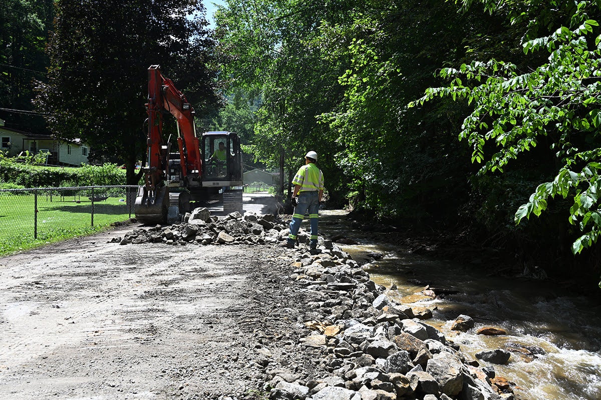 An excavator and a highway worker in a high-visibility vest rebuild a dirt road damaged by floods, reinforcing the bank alongside a flowing creek with large stones