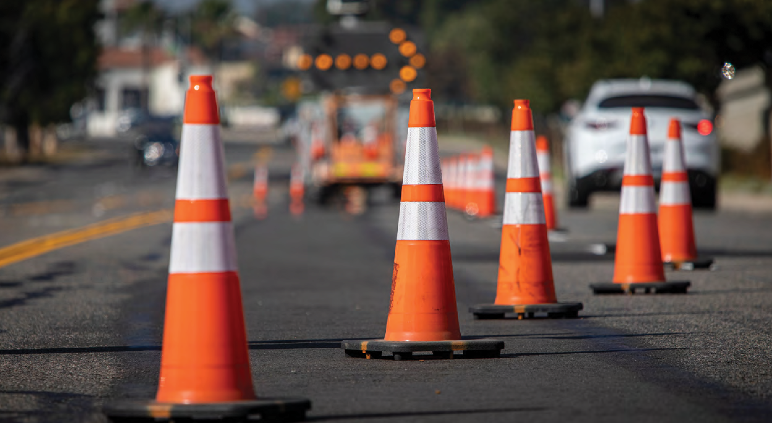 Row of bright orange traffic cones with reflective white stripes setting up a lane closure on a paved road.