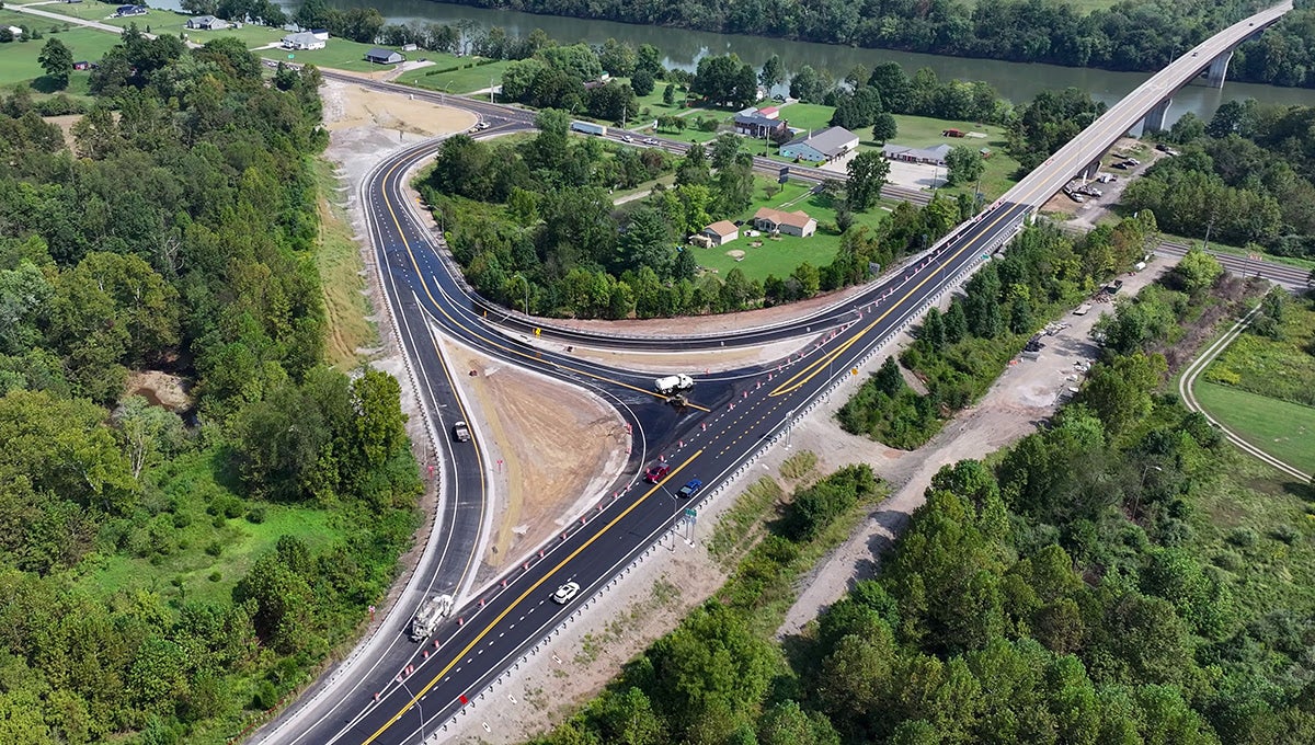 Aerial view of a newly paved highway interchange surrounded by trees, showing fresh asphalt, painted road markings, connected ramps, and a long bridge spanning a river in the background.